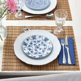 Table set with white plates featuring blue floral patterns, bamboo placemats, silverware, and glasses.