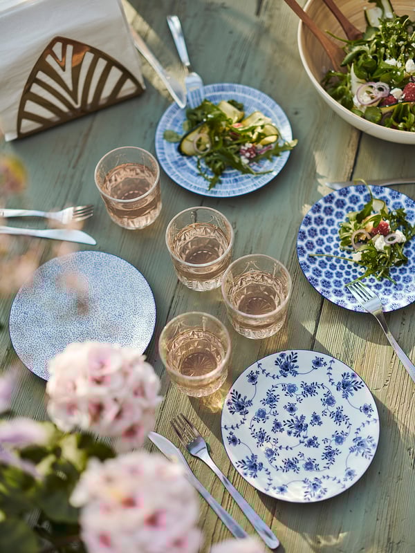 A wooden table set with blue floral patterned ENTUSIASM plates, glasses, and utensils. Pink flowers decorate the scene, adding charm.