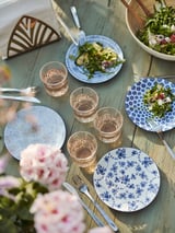 A wooden table set with blue floral patterned ENTUSIASM plates, glasses, and utensils. Pink flowers decorate the scene, adding charm.