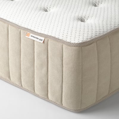 Image shows a corner view of a high-quality mattress with visible stitching and labels, indicating its brand and materials.