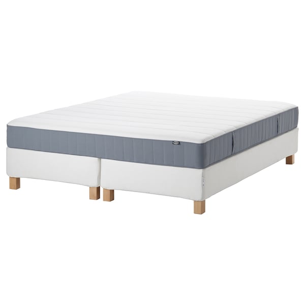 A white and blue mattress on a bed frame. The mattress has a padded surface and a visible spring system. The bed frame has light wooden legs.