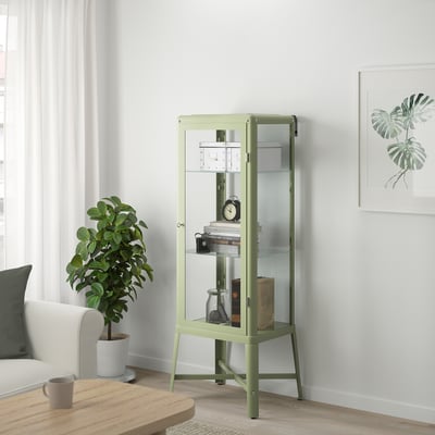 Fabriker cabinet in green, tall, glass-door, fits collectibles, stands near white couch, green plant, wooden table.