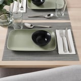A dining setup with FÄRGKLAR bowl on rectangular green plate, cutlery, and glass on gray placemat.