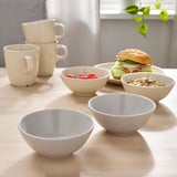Four white bowls and three mugs arranged on a wooden table, containing food items such as berries, cereal, and a sandwich.
