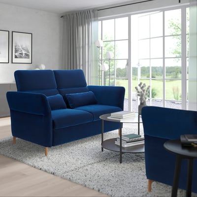 Modern minimalist living room with large blue FAMMARP sofa by window, blue armchair, glass coffee table, rug, and large window offering outdoor view.