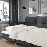Modern gray sofa-bed with white bedding, adjustable headrests and armrests in a cozy room with wall art, floor lamp, and plant.