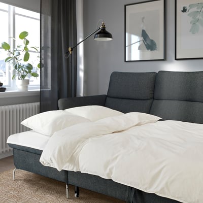 Modern gray sofa-bed with white bedding, adjustable headrests and armrests in a cozy room with wall art, floor lamp, and plant.