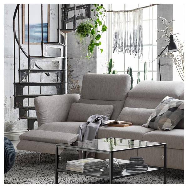 Modern grey sofa with adjustable headrests & armrests, near metal spiral staircase, glass coffee table, and large window with hanging plants.