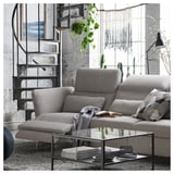 Modern grey sofa with adjustable headrests & armrests, near metal spiral staircase, glass coffee table, and large window with hanging plants.