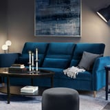 Modern living room with teal FAMMARP sofa, gray ottoman, dark coffee table with candles, books, and magazines. Abstract blue artwork on wall.