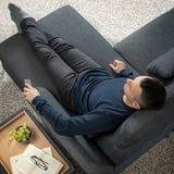 A man relaxes on a dark grey FAMMARP sofa, holding a remote, with grapes and a book beside him.