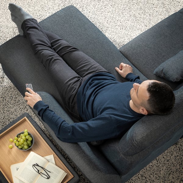 Man reclines on FAMMARP sofa, remote in hand, beside grapes and book.