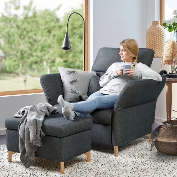 Woman relaxes in modern gray FAMMARP armchair, sipping coffee, with large window and green view.