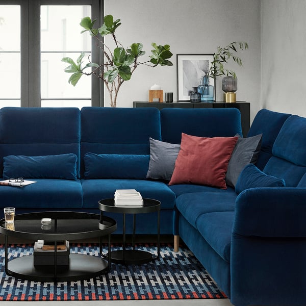 Modern living room with blue velvet sofa, colorful cushions, patterned rug, glass tables, plants, and wall decor.