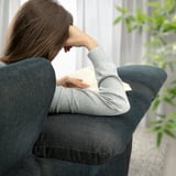 A person sitting on a dark green FAMMARP sofa, reading a book near a window with plants.