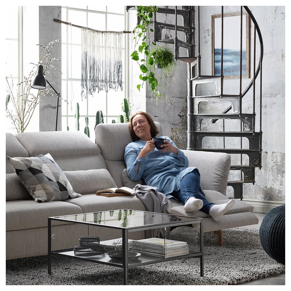 A woman on a beige FAMMARP sofa, using a tablet, with a spiral staircase and large window in a modern, industrial-style room.