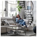 A woman on a beige FAMMARP sofa, using a tablet, with a spiral staircase and large window in a modern, industrial-style room.