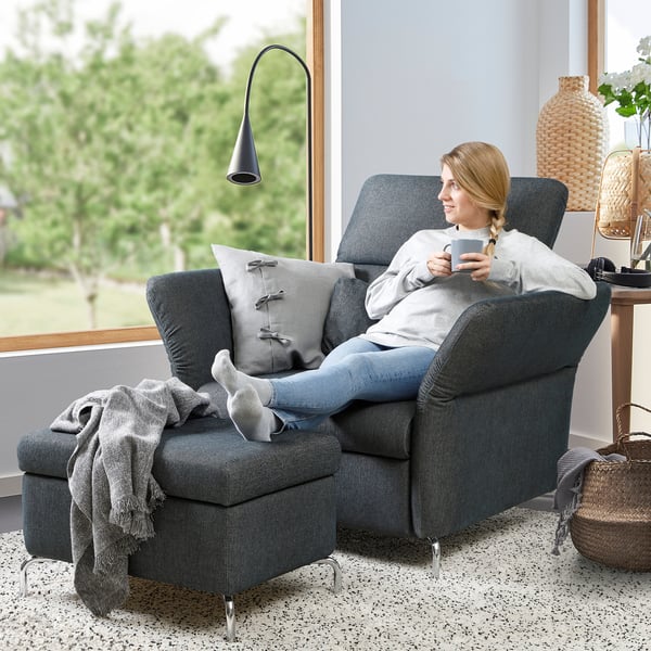 Woman relaxes in grey FAMMARP reclining sofa, holding a mug, with footrest extended and grey throw pillows, near a large window.