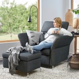 Woman relaxes in grey FAMMARP reclining sofa, holding a mug, with footrest extended and grey throw pillows, near a large window.