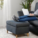 Person sitting on blue FAMMARP sofa, feet up, using a laptop. Sofa has adjustable headrests and wooden legs. Green plant in background.