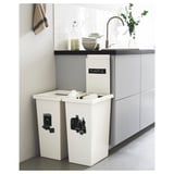 Modern kitchen scene with two white FILUR recycling bins labeled plastic and glass, positioned under a counter with a sink and a potted plant.