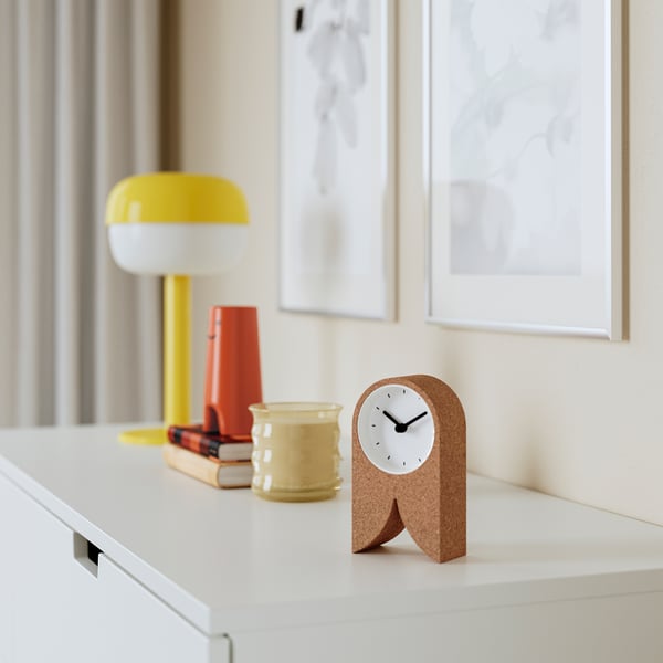 Small FLODKRÄFTA cork clock sits quietly on a bright desk; yellow lamp, cup, & books nearby.