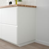 FÖRBÄTTRA white kitchen cabinet, sleek and modern design, tall with a wide base, light-colored wooden countertop, small decorative items on top.