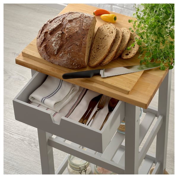 A FÖRHÖJA wooden kitchen cart with a grey drawer. It has a cutting board with sliced bread and a knife. The drawer holds napkins and cutlery.