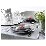 Elegant dining setup with gray plates, silver cutlery, glassware, and decorative wrapped napkins. White flowers in a clear vase as centerpiece.