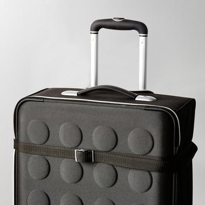 Black suitcase with raised circles, pull handle up, ready to travel. Strap and buckle visible, securing contents.