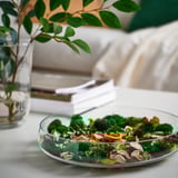 Glass bowl: clear, round, holding green and brown dried plants, small, near books and vase.