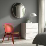 Modern bedroom with red FRÖSET chair, white dresser, plant, and round mirror. Light gray walls. Bed partially visible. Efficient space use.