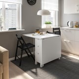 Small, white, foldable dining table with two black chairs in modern kitchen. Space-saving design, positioned near window and cabinet.