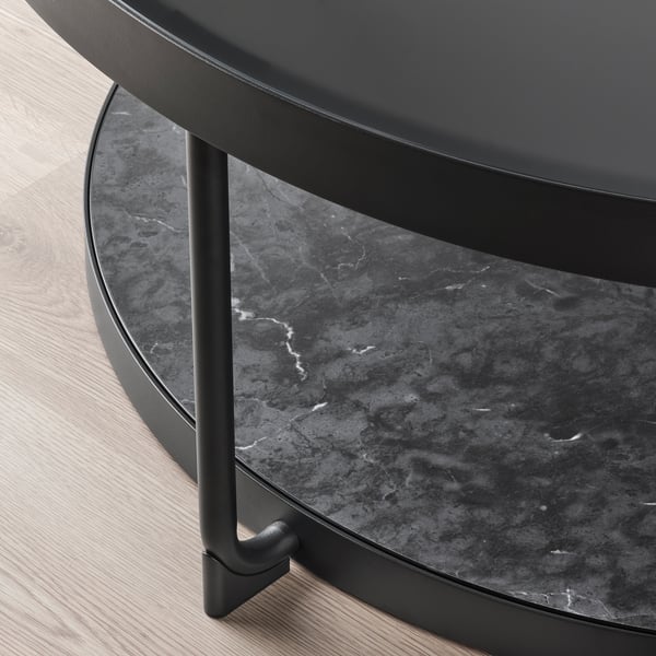 Modern round side table with sleek black design, featuring tempered glass top and metallic legs, sits on wooden floor.