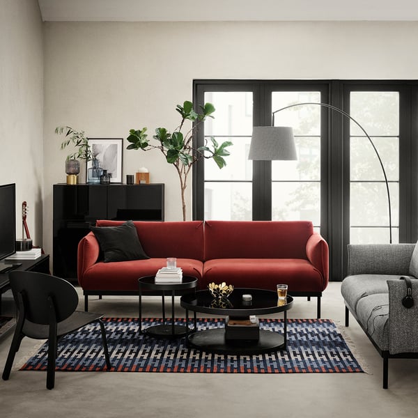 Modern living room with red l-shaped couch, grey armchair, round black glass side tables, colorful rug, and large windows allowing natural light.