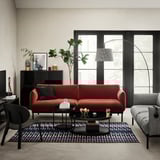 Modern living room with red l-shaped couch, grey armchair, round black glass side tables, colorful rug, and large windows allowing natural light.