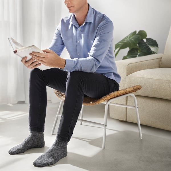Man sitting on woven stool, reading book. Light-filled room with sofa, plant, and window.