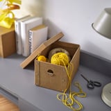 Small yellow yarn balls in brown cardboard box, on gray desk with books and scissors.
