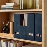 Wooden bookshelf with dark blue velvet magazine holders storing papers and a black vase with pens.