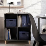 Modern, minimalist desk setup with GJÄTTA black cube storage, containing blue velvet boxes, organized documents, and a sleek lamp.
