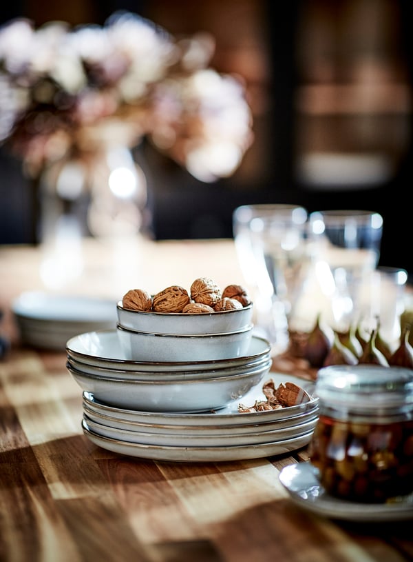 Stacked white ceramic dishes, including bowls and plates, on wooden table. Walnuts in top bowl, glasses, and jar nearby.