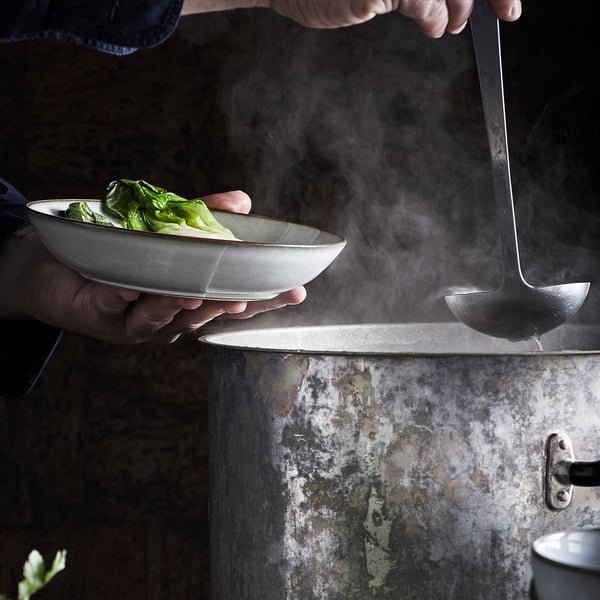 Hands holding glazed bowl with green veggies over steamer.