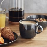 Dark grey mug, orange juice, croissant, and granola on a wooden table, showcasing a stylish breakfast setup.