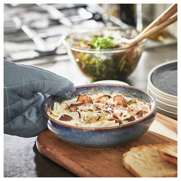 A hand grabs a blue glazed ceramic dish filled with baked food, next to a salad bowl and bread on a wooden board.