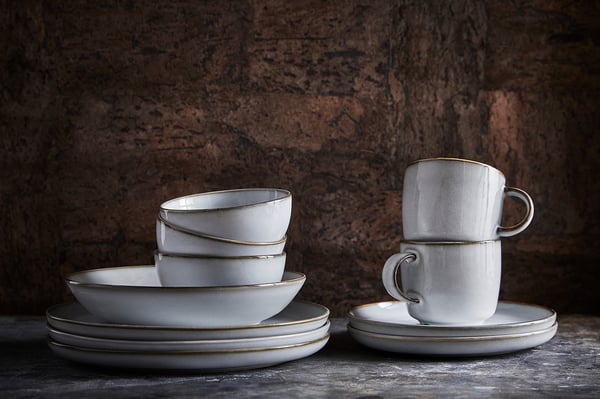 White GLADELIG stoneware tableware, including plates and cups, displayed on a grey surface with a dark brick wall background.