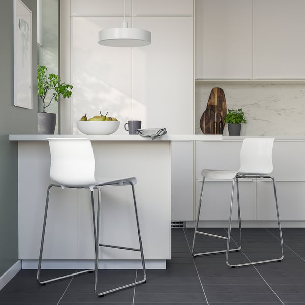 Modern kitchen with white GLENN stools. Sleek chrome legs, comfortable white seats. Hanging light, potted plants, fruits on counter.