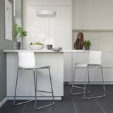 Modern kitchen with white GLENN stools. Sleek chrome legs, comfortable white seats. Hanging light, potted plants, fruits on counter.