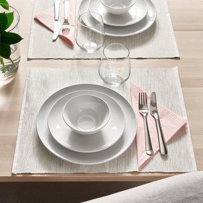 Two table settings with white plates, bowls, glasses, and pink napkins on woven placemats, ready for dining.