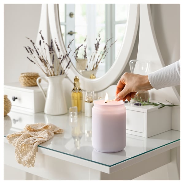 A person lights a lavender-colored GOLIATTUJA scented candle on a glass shelf. The candle is cylindrical, and the setting includes a mirror and decor.
