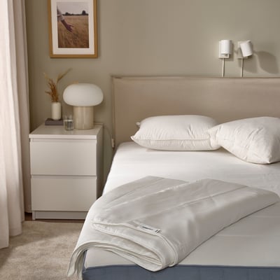 Bed with beige GRÄSROTSFLY duvet folded at end, white pillows, nightstand with lamp and books. Wall with picture frame. Cozy, minimalist bedroom.